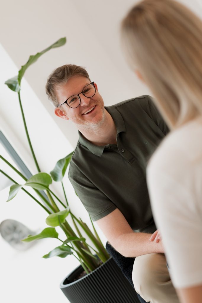 Beratungsgespräch Mann mit Brille sitzt lächelnd neben einer Pflanze, Blickrichtung auf eine Person.